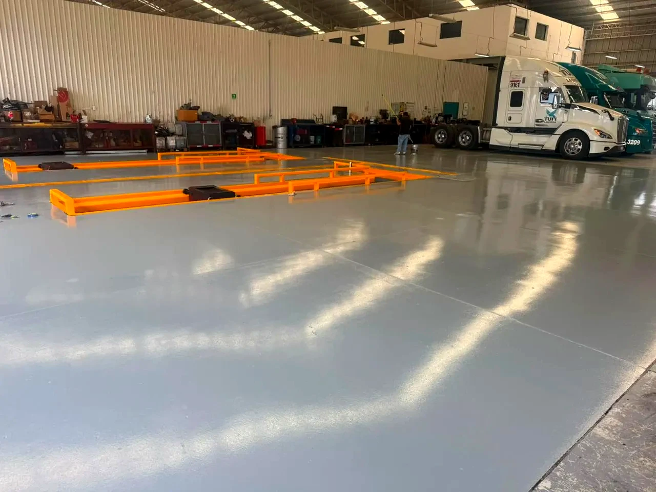 Piso gris terminado en el taller de mantenimiento, con bordes de fosas en amarillo y tractocamiones al fondo